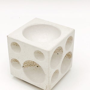 May include: White concrete cube planter with multiple circular cutouts. The planter has a large circular cutout on the top.
