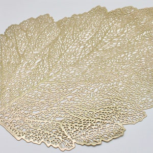 Könnte beinhalten: Goldfarbener Platzdeckchen in Blattform mit einem filigranen, komplizierten Design. Der Platzdeckchen ist aus einem dünnen, metallischen Material gefertigt und hat eine leicht erhabene Textur.