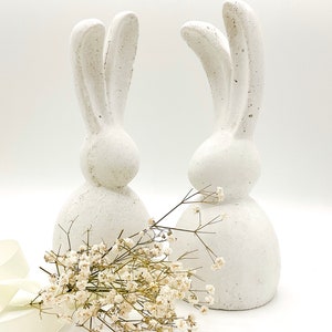 May include: Two white ceramic bunny figurines with long ears, standing on a white surface with a bouquet of white baby's breath flowers.