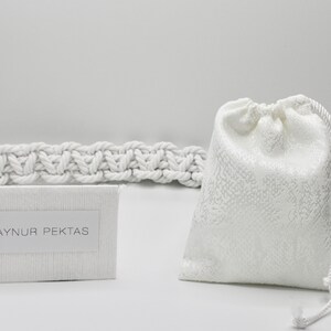 May include: White drawstring bag with a white rope handle. A white card with the text "AYNUR PEKTAS" is in the foreground. A white rope is in the background.