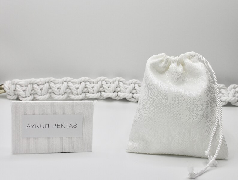 May include: White drawstring bag with a white rope handle. A white card with the text "AYNUR PEKTAS" is in the foreground. A white rope is in the background.