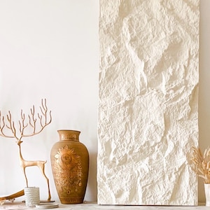May include: A decorative display featuring a large, textured, off-white wall panel. A bronze-colored deer sculpture and a detailed vase with a sunburst design are on a white cabinet. Dried plants in a small vase add a natural touch.
