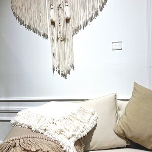 May include: A white couch with two pillows, one beige and one white, with a white fringed throw blanket draped over the arm. A white macrame wall hanging with fringe is hanging on the wall behind the couch.