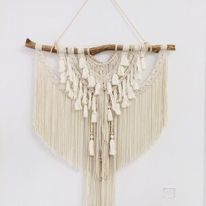 Könnte beinhalten: Ein großer, weißer Makramee-Wandbehang mit einem natürlichen Ast oben. Der Behang verfügt über eine Vielzahl von Knoten und Quasten, die eine  boho-moderne Ästhetik schaffen.