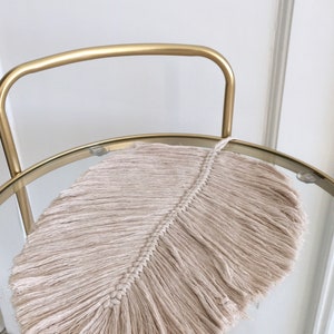 Könnte beinhalten: Eine beige Makramee-Feder mit langen Fransen, die auf einem goldenen Metall- und Glastisch liegt.
