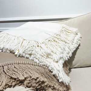 May include: Two throw pillows with fringe trim. The top pillow is white and the bottom pillow is a light brown.