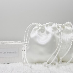 May include: Two white satin drawstring bags with white cords. The bags are sitting on a white surface. The text "Anya Life Presents" is printed on a white card in front of the bags.