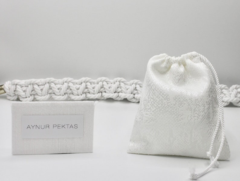 May include: A white drawstring bag with a white rope handle. The bag is sitting on a white surface next to a white card with the text "AYNUR PEKTAS" printed on it. The card is sitting on top of a white rope.