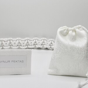 May include: A white drawstring bag with a white rope handle. The bag is sitting on a white surface next to a white card with the text "AYNUR PEKTAS" printed on it. The card is sitting on top of a white rope.