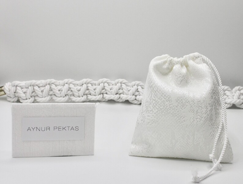 May include: A white drawstring bag with a white rope handle. The bag is sitting on a white surface next to a white card with the text "AYNUR PEKTAS" printed on it. The card is sitting on top of a white rope.