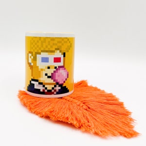 Könnte beinhalten: Eine gelbe Keramiktasse mit einem Pixelbild einer Person, die eine 3D-Brille trägt und eine rosa Blase bläst. Die Tasse steht auf einem orangefarbenen Untersetzer mit Fransen.