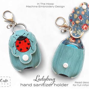 以下が含まれることがあります： 水色のハンドサニタイザーホルダー。赤いテントウムシのデザインで、黒い斑点と触角があります。ホルダーは布製で、金色のクリップと白いボタン留めが付いています。「Ladybug hand sanitizer holder」の文字が見えます。