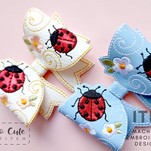 Puede incluir: Dos lazos de pelo de tela bordados con un diseño de mariquita. Los lazos son blancos y azul claro con costuras amarillas. La mariquita es roja con manchas negras y detalles blancos. El texto "So Cute Stitch" es visible en la esquina inferior izquierda. El texto "ITH Machine Embroidery Design" es visible en la esquina inferior derecha.