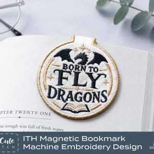 Könnte beinhalten: Ein kreisförmiges magnetisches Lesezeichen aus weißem Stoff mit goldener und schwarzer Stickerei. Das Design zeigt eine schwarze Drachensilhouette, Sterne, ein offenes Buchsymbol und den Text BORN TO FLY DRAGONS in schwarzer Schrift mit Goldrand.