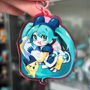 May include: A pink-edged acrylic keyring featuring a cartoon character with teal hair and a blue outfit. The character holds a circular object and is with a small yellow creature. The keyring is held by a pink ring.