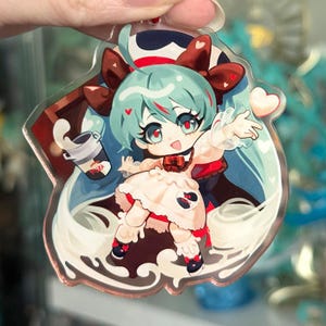 May include: A clear acrylic charm depicting a cartoon character with teal hair, red eyes, and a white dress. The character holds a milk cup and has a heart detail. The charm has a brown and white background.