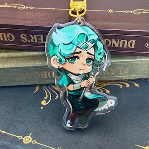 Peut inclure: Un porte-clés en acrylique transparent représentant un personnage de dessin animé aux cheveux turquoise et aux vêtements turquoise et noirs. Le personnage tient un bâton et a une expression triste. Le porte-clés a un fermoir doré.