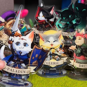 Puede incluir: Colección de figuras de gatos de acrílico, cada una representando una clase de fantasía diferente. El gato Paladín sostiene una espada y un escudo, mientras que el gato Clérigo tiene un emblema solar. Otras clases incluyen Brujo y Druida. Las figuras están sobre bases transparentes.