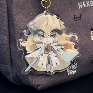 K&ouml;nnte beinhalten: Ein durchsichtiger Acryl-Schl&uuml;sselanh&auml;nger mit einer Comicfigur mit blonden Haaren, die ein wei&szlig;-goldenes Outfit tr&auml;gt und ein Schwert h&auml;lt. Die Figur hat H&ouml;rner und blaue Augen.