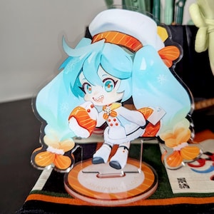 May include: A clear acrylic figure of an anime-style character with blue hair, wearing a white outfit with orange accents. The figure is holding a sushi roll and stands on a round base with text. The character has a cheerful expression.