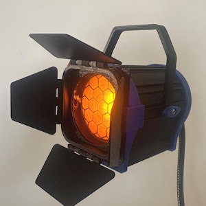 Puede incluir: Un foco de escenario negro y azul con viseras metálicas ajustables y una lente central con un patrón de rejilla hexagonal visible. La luz emite un brillo naranja cálido desde el interior de la carcasa, que incluye un asa superior.