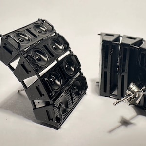 May include: A pair of black speaker earrings. The earrings are shaped like small speaker stacks with multiple speakers on each side. The earrings are made of metal and have a silver post.