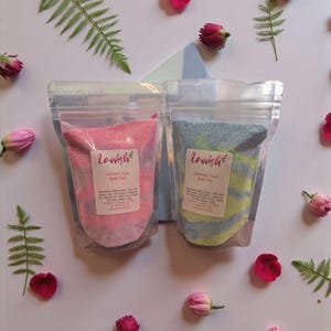 Puede incluir: Dos bolsas transparentes de sales de baño, una rosa y otra azul, etiquetadas como "Lavish". La bolsa rosa es "Unicorn Dust", la azul es "Dinosaur Dust". La imagen está decorada con flores rosas, hojas de helecho y una forma de papel azul claro y blanco.