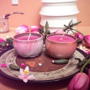 May include: Two pink candles in ceramic bowls sit on a decorative plate, surrounded by pink tulips and petals. The candles have white wicks and are set against a peach-colored background. A stack of white bowls is in the background.