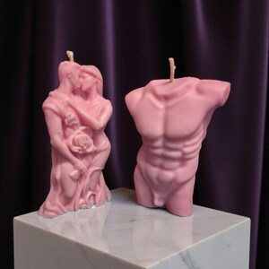 Puede incluir: Dos velas rosas. Una vela representa una pareja abrazándose y besándose, sosteniendo una rosa. La otra vela es un torso esculpido. Ambas velas están sobre un cubo de mármol blanco, sobre un fondo púrpura.