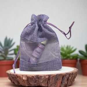 May include: A small, lavender drawstring bag with a sheer mesh front, containing a small essential oil bottle. The bottle is labelled "Sleep." The bag sits on a wooden slice, with potted succulents in the background.