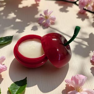 May include: A cherry-shaped lip balm container, red with a green stem, open to reveal a white balm. The background features pink cherry blossoms and green leaves, creating a natural, springtime aesthetic.