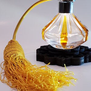 May include: A clear glass perfume bottle with a gold tassel and a black wooden base. The bottle has a diamond-shaped design and a silver atomizer.