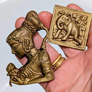 May include: Two antique brass figurines. One is a detailed profile of a woman with ornate hair and clothing, holding a flower. The other is a square plaque with a relief of an elephant. Both are held in a hand.