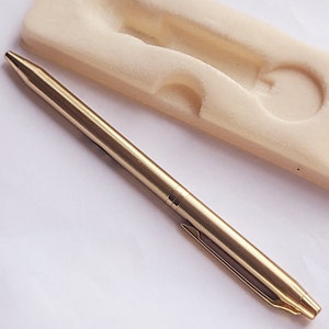 May include: A gold-colored pen with a textured, ridged design. The pen is lying next to a beige, molded foam insert, likely from a pen case. The pen has a clip and a pointed tip.