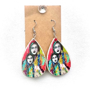 May include: Teardrop-shaped earrings with a silver-colored border. Each earring features a colorful illustration of two women with long braids and a red accent at the top. The background is a mix of red, yellow, and blue.