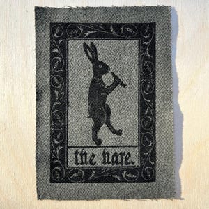 May include: Black and white illustration of a rabbit playing a flute, framed in a decorative border with the text "the hare".