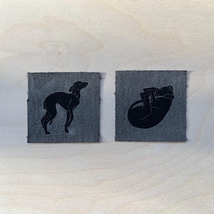 Könnte beinhalten: Zwei Denim-Patches mit schwarzen Hundesilhouetten. Ein Patch zeigt einen stehenden Hund, der andere einen zusammengerollten Hund. Die quadratischen Patches haben eine strukturierte Optik, vor einem hellen Holzgrund.