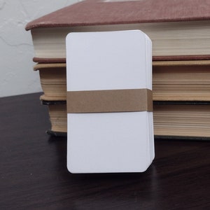 May include: A stack of three books with a white blank card with a brown band on top. The books are brown, tan, and red.