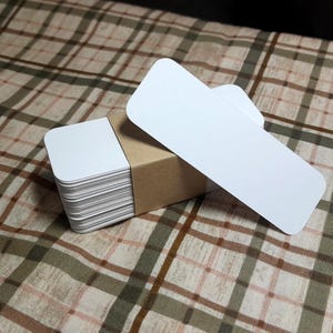 Puede incluir: Un mont&oacute;n de tarjetas rectangulares blancas con esquinas redondeadas, sujetas por una banda de papel marr&oacute;n. Dos tarjetas adicionales se colocan encima. Las tarjetas est&aacute;n sobre una superficie con estampado de cuadros marrones y verdes.