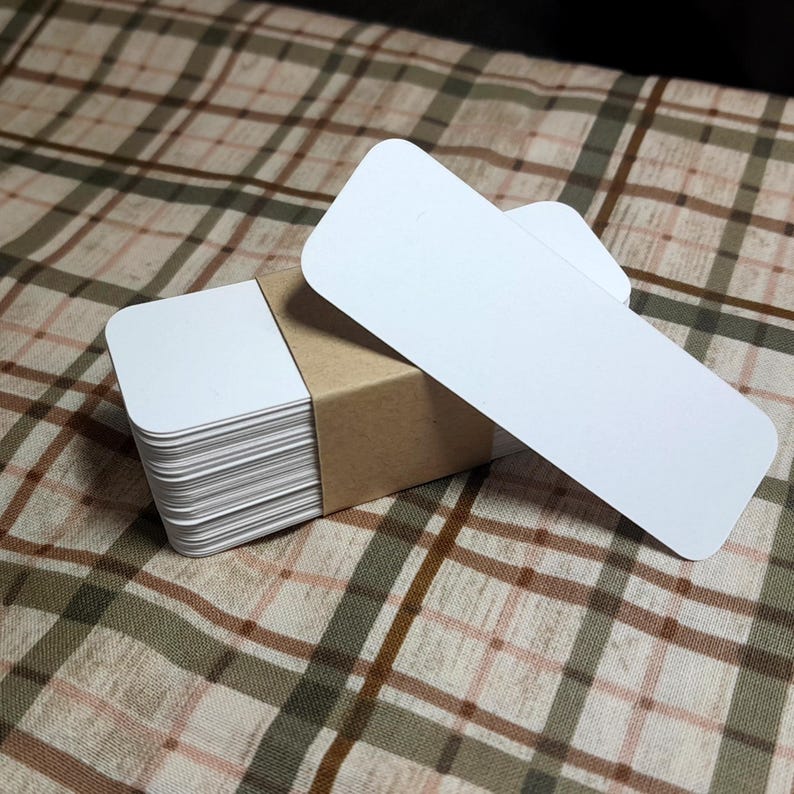 Puede incluir: Un mont&oacute;n de tarjetas rectangulares blancas con esquinas redondeadas, sujetas por una banda de papel marr&oacute;n. Dos tarjetas adicionales se colocan encima. Las tarjetas est&aacute;n sobre una superficie con estampado de cuadros marrones y verdes.