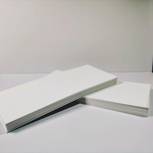 May include: Two stacks of rectangular white paper sheets are positioned on a flat surface. The larger stack is oriented horizontally on the left, while a smaller, square-shaped stack of white paper is placed to the right of the first stack.