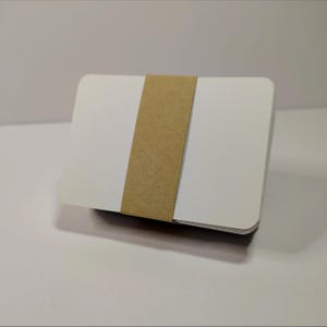 May include: A stack of rectangular white cards with rounded corners, secured together by a wide, textured light brown paper band that wraps vertically around the centre of the cards.