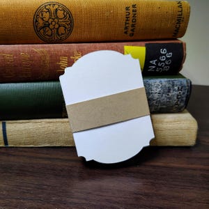 May include: A stack of white paper tags with decorative scalloped edges, secured together by a plain brown kraft paper band. The tags feature a smooth, matte surface and a symmetrical, rounded shape with notched corners at the top and bottom.