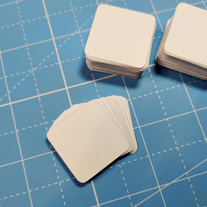May include: Three stacks of blank, off-white square paper cards with rounded corners. The cards are arranged in two upright piles and one fanned-out pile, all featuring a smooth, matte surface texture and uniform edges throughout the collection.