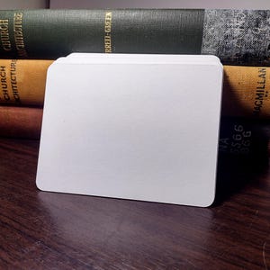 May include: A stack of blank, white, rounded-corner cards. The cards are stacked neatly on a dark wooden surface, with the spines of several antique books in the background. The books are various colors, including green and tan.