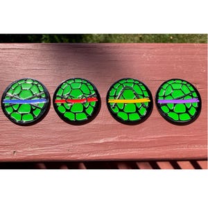 May include: Four round, green Teenage Mutant Ninja Turtles-themed coasters. Each coaster features a different colored band and weapon design: blue, red, yellow, and purple. The coasters have a black border and a textured surface.