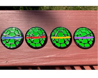 Pode incluir: Quatro bases para copos redondas com o tema Tartarugas Ninja. Cada base apresenta um design de carapaça de tartaruga verde com uma faixa e uma arma de cor diferente: azul, vermelho, amarelo e roxo. Borda preta.