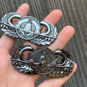 May include: Two metal military insignia badges. One is silver-toned, the other is black. Each features a central emblem with a winged design and a parachute. The badges are detailed with intricate patterns and textures.