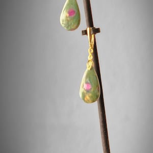 May include: Gold-coloured earrings featuring teardrop-shaped green and pink stones. The earrings are suspended from gold-coloured lever-back closures. The stones have a marbled appearance, with a mix of green, pink, and gold hues.
