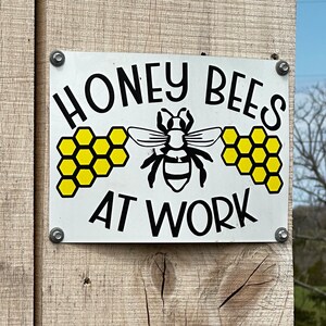 May include: A rectangular white metal sign with black text that reads HONEY BEES AT WORK. The sign features a central black illustration of a bee flanked by two yellow honeycomb patterns on either side, secured with four silver screws to a wooden post.